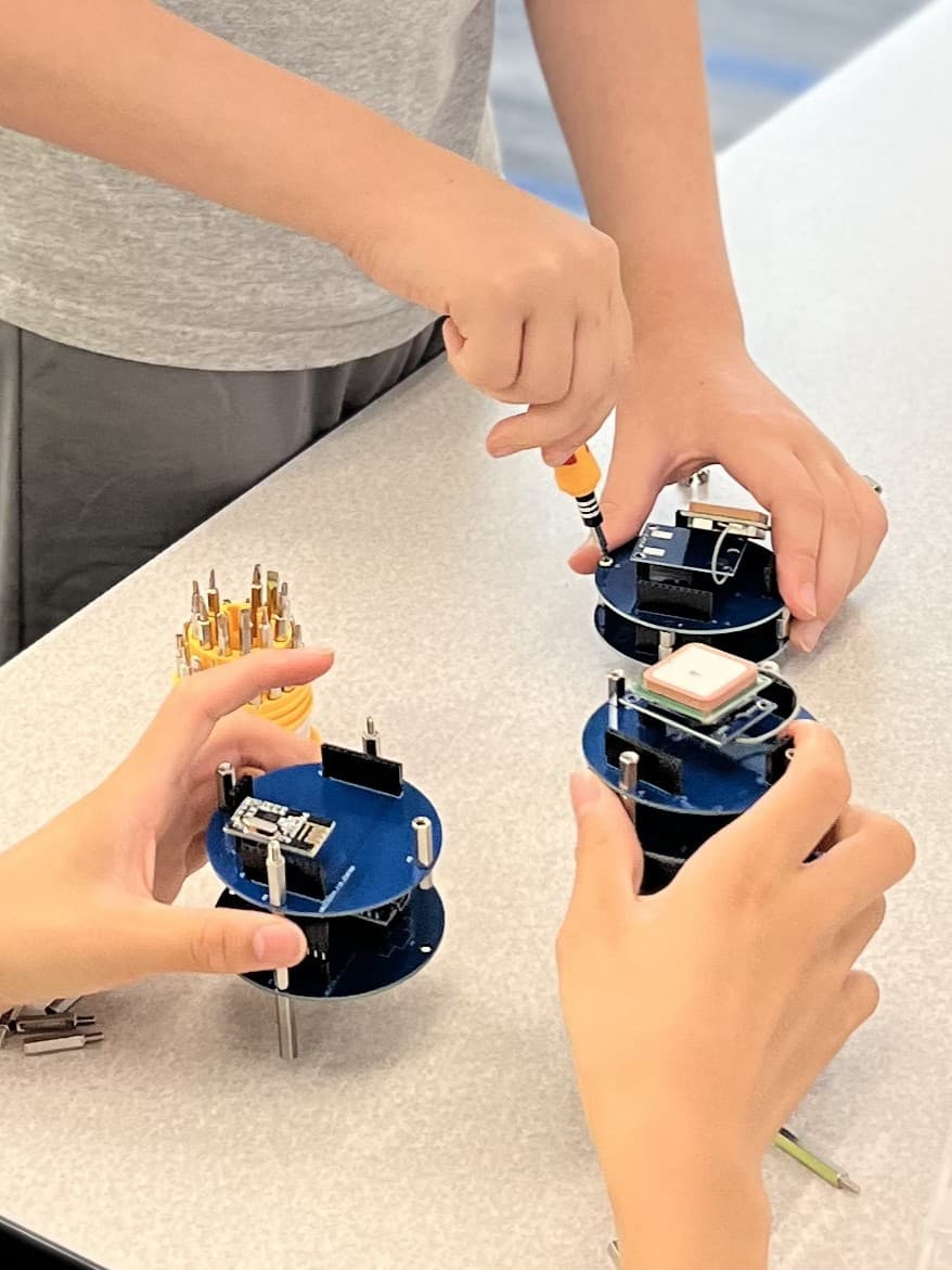 Students assembling the stacked circuit-board decks of a CanSat satellite, one holding a GPS patch antenna in place while another drives a screw.